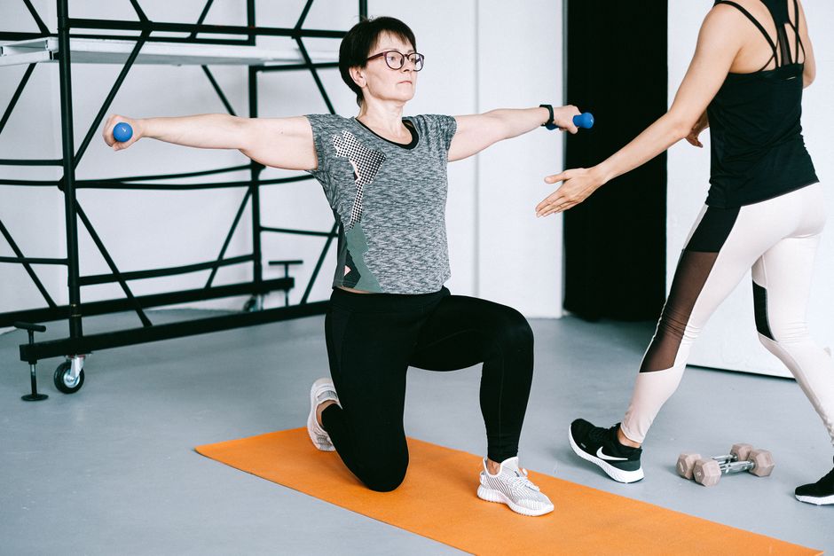 Two women working out with dumbbells on yoga mats in a modern gym setting, focusing on strength training.