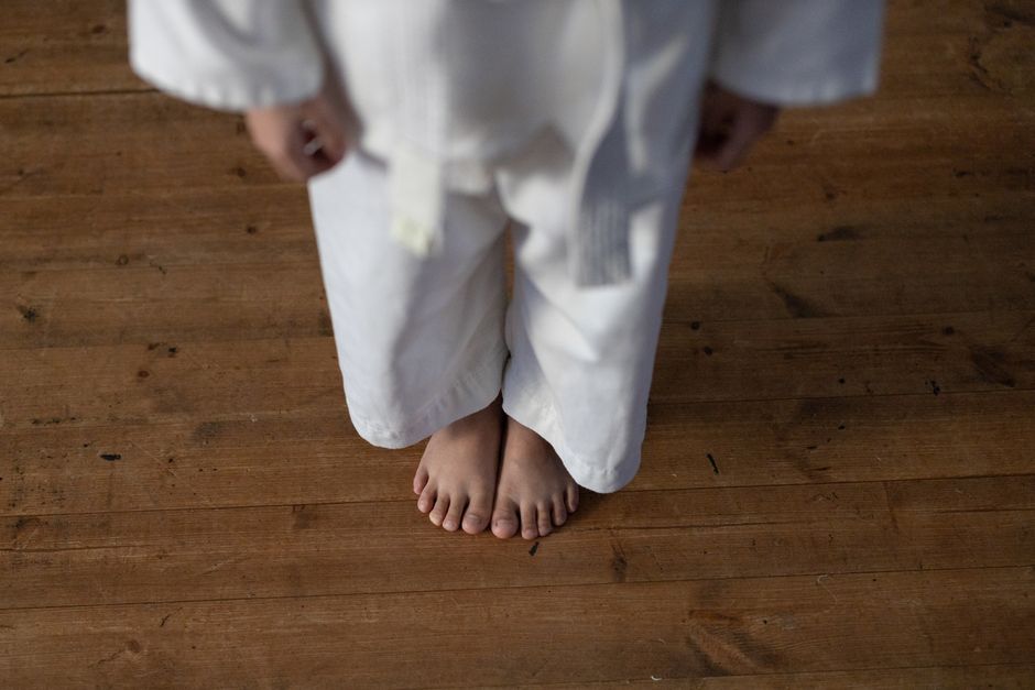 A child standing barefoot on a wooden floor wearing a white karate uniform and belt.