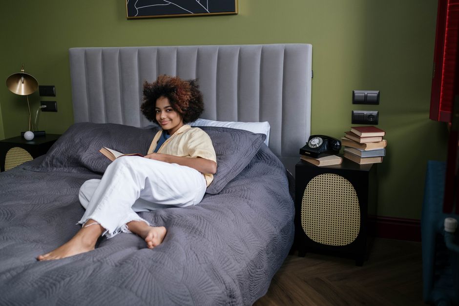 A woman with afro hair lying on a bed reading a book, depicting a cozy indoor setting.