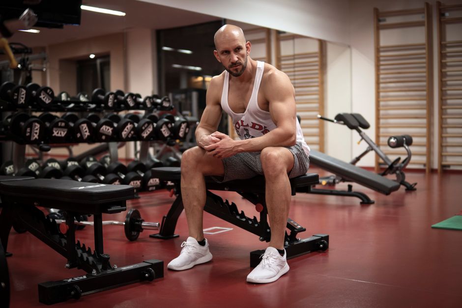 Strong, determined man in white tank top sitting in gym, showcasing fitness and strength.