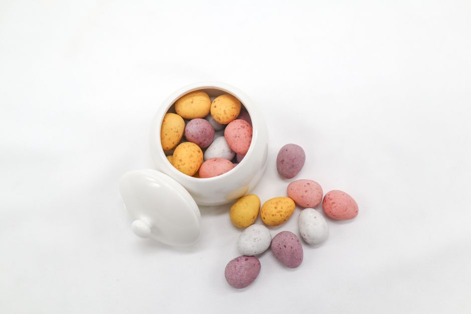 An overhead view of colorful candy eggs in a white ceramic dish on a white background.