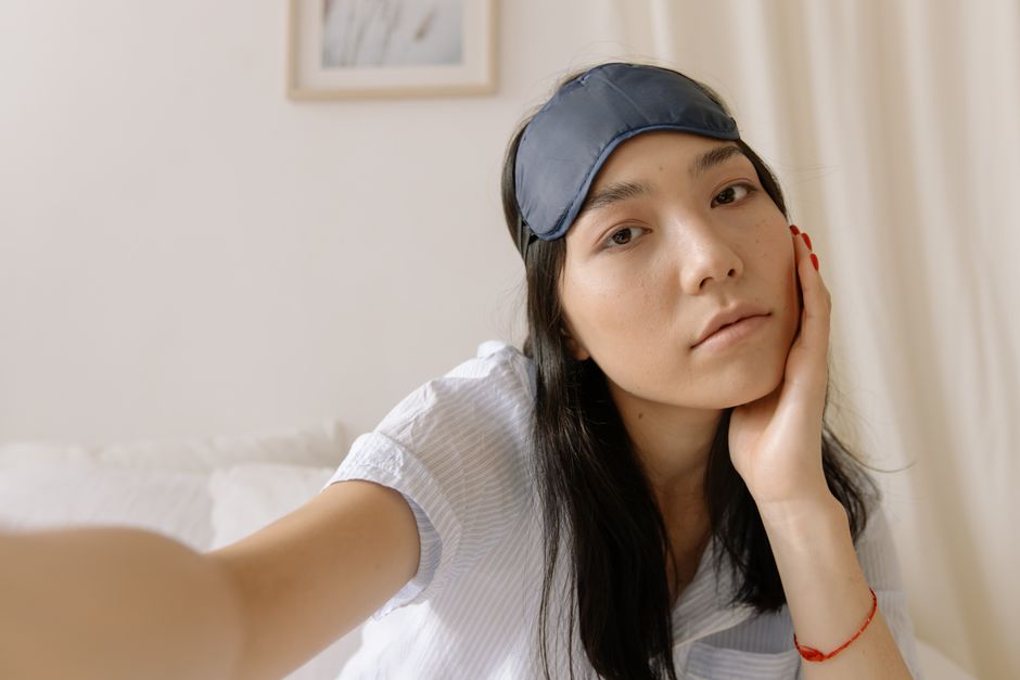Close-up of a young woman indoors wearing a sleep mask, resting her head on her hand.
