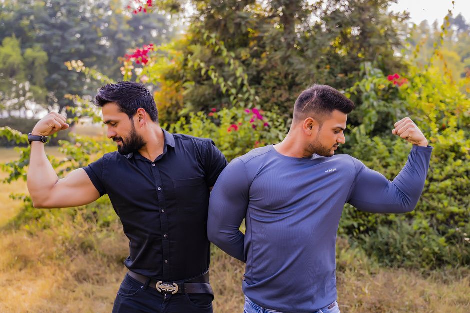 Two muscular men flexing their biceps outdoors in a garden setting.