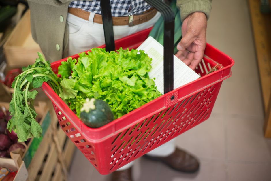 Close-up of a hand carrying a shopping basket filled with fresh greens in a market.
