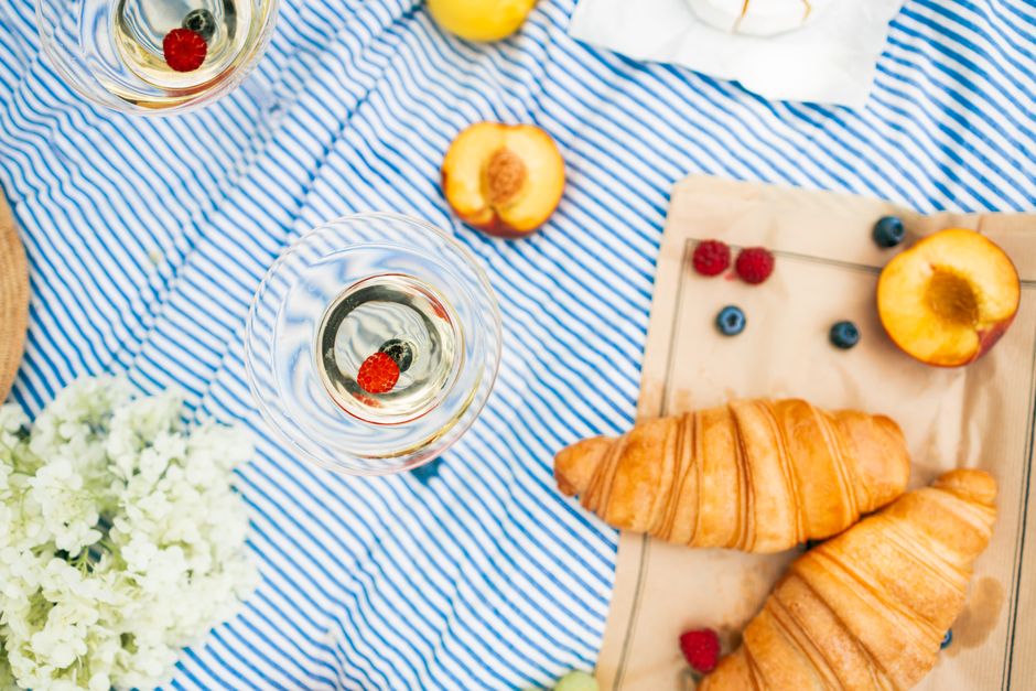 Overhead view of a picnic with croissants, wine, and fresh fruit on a striped blanket.