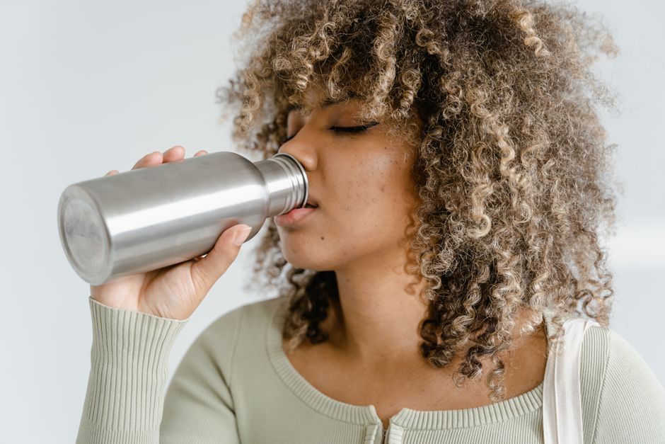 Stylish woman with curly hair drinking water from a stainless steel bottle indoors.