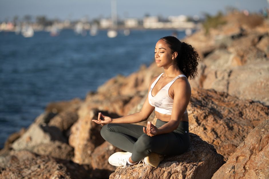 Woman meditating on rocks by the ocean, embodying calmness and wellness.