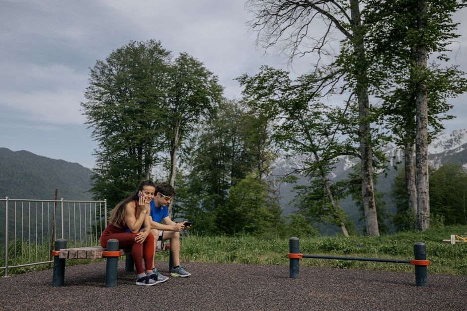 A couple in sports clothing sitting on a bench outdoors, surrounded by trees and mountains.