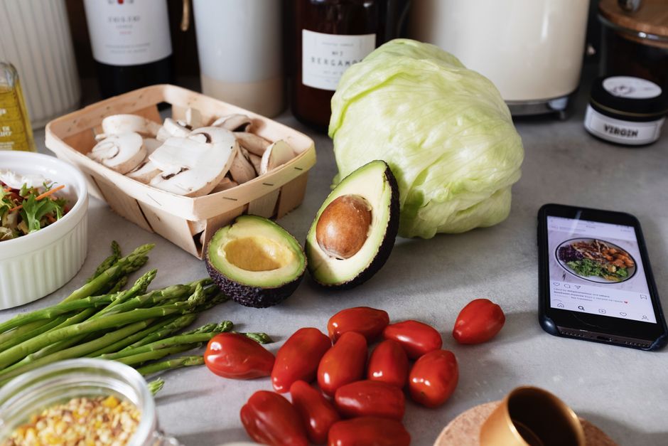 Fresh organic chopped vegetables placed on countertop near kitchen utensils and mobile phone in bright kitchen during preparation salad according to recipe from smartphone