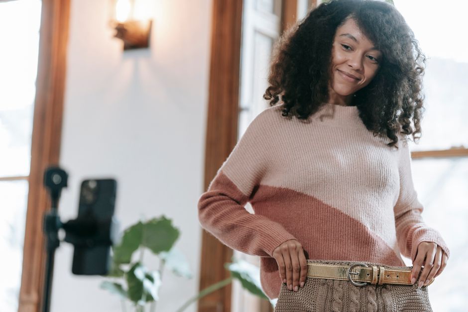 Cheerful woman with curly hair wearing warm sweater trying on knitted beige skirt and making selfie photo on smartphone fixed on tripod