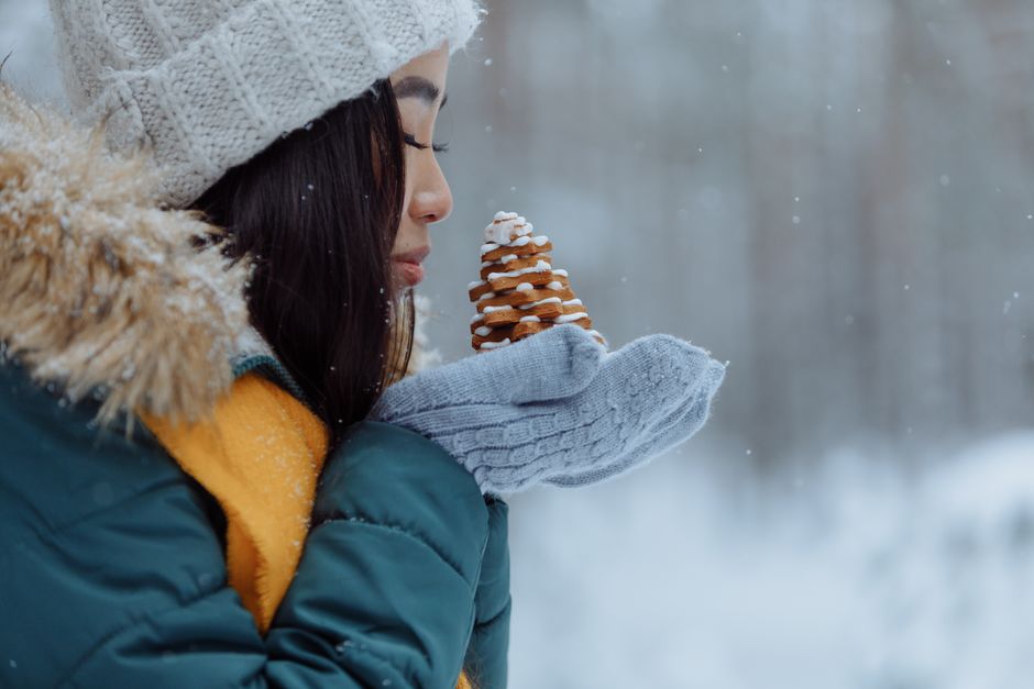 Asian woman in a winter coat holds Christmas cookies in a snowy landscape.